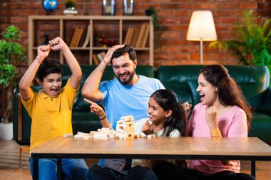 Hintli ebeveynler küçük çocuklarla vakit geçirmenin keyfini çıkarırken ve rahat modern oturma odasında ahşap blokları dikkatlice dengelerken aile evde ahşap blok oyunu oynuyor.