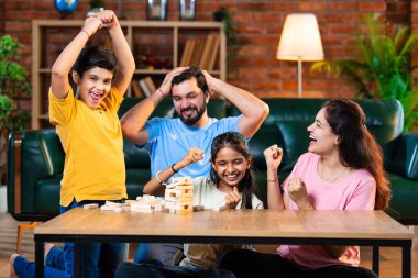 Hintli ebeveynler küçük çocuklarla vakit geçirmenin keyfini çıkarırken ve rahat modern oturma odasında ahşap blokları dikkatlice dengelerken aile evde ahşap blok oyunu oynuyor.