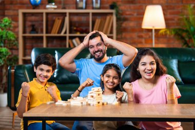 Hintli ebeveynler küçük çocuklarla vakit geçirmenin keyfini çıkarırken ve rahat modern oturma odasında ahşap blokları dikkatlice dengelerken aile evde ahşap blok oyunu oynuyor.