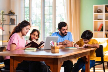 Ebeveynler evde çocuklara bilgisayar ve kitap öğretiyor, Hintli baba ve anne, oğullarına ev ödevlerinde, okumada, yazmada ve online çalışmada yardım ediyorlar.