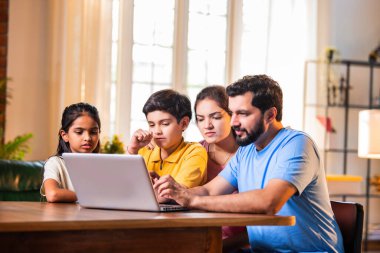 Aile evde dizüstü bilgisayar kullanıyor modern oturma odasındaki yemek masasında oturuyor, Hintli ebeveynler çocuklar kayıyor, sörf yapıyor, tatil planlıyor ya da internetten alışveriş yapıyor ve kaliteli zaman geçiriyor.