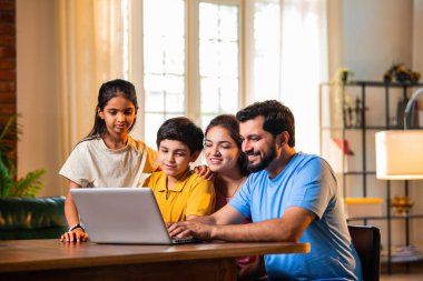 Aile evde dizüstü bilgisayar kullanıyor modern oturma odasındaki yemek masasında oturuyor, Hintli ebeveynler çocuklar kayıyor, sörf yapıyor, tatil planlıyor ya da internetten alışveriş yapıyor ve kaliteli zaman geçiriyor.