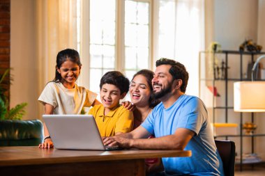 Konforlu bir evde yemek masasında otururken dizüstü bilgisayardan komik filmler izlemek Hintli ebeveynler ve çocuklar yüksek sesle gülüyor. Aile şovu, komedi videosu, güçlü bir bağ ve kaliteli aile zamanı paylaşımı.