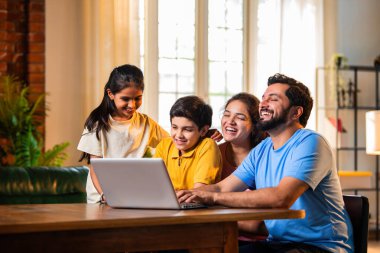 Konforlu bir evde yemek masasında otururken dizüstü bilgisayardan komik filmler izlemek Hintli ebeveynler ve çocuklar yüksek sesle gülüyor. Aile şovu, komedi videosu, güçlü bir bağ ve kaliteli aile zamanı paylaşımı.