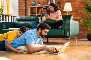 Baba oğul çalışırken Hintli adam dizüstü bilgisayarı yerde yatarken ve çocuğu üzerinde kestirirken, anne ve kızı rahat modern oturma odasında akıllı telefon kullanarak koltukta otururken, baba çalışıyor.