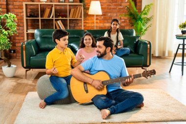 Babam gülen Hintli eş, oğul ve kızıyla gitar çalıyor. Müsrif oturma odasında oturuyor. Müzikal kaynaşmanın ve modern evde kaliteli zaman geçirmenin tadını çıkarıyor.