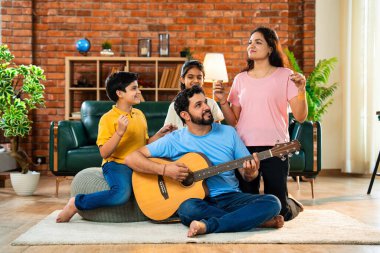 Babam gülen Hintli eş, oğul ve kızıyla gitar çalıyor. Müsrif oturma odasında oturuyor. Müzikal kaynaşmanın ve modern evde kaliteli zaman geçirmenin tadını çıkarıyor.