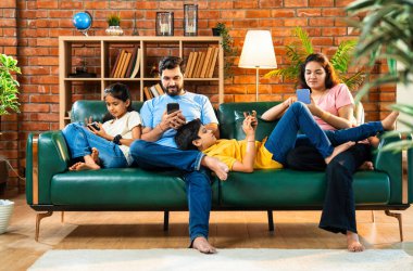 Hintli aile, oturma odasındaki kanepede tek başına otururken akıllı telefonları kullanıyor. Ekran bağımlılığını modern yaşam tarzında gösteriyorlar. Etkileşim ya da bağ kurma yok.