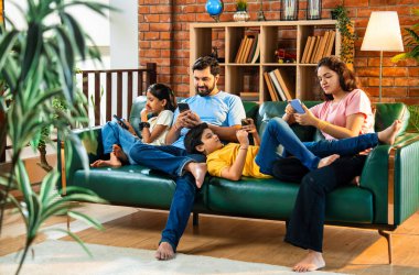Hintli aile, oturma odasındaki kanepede tek başına otururken akıllı telefonları kullanıyor. Ekran bağımlılığını modern yaşam tarzında gösteriyorlar. Etkileşim ya da bağ kurma yok.