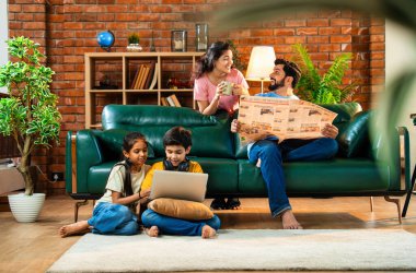 Çocuklar dizüstü bilgisayar kullanıyor, babaları gazete okuyor aileleriyle rahat bir koltukta oturuyor, kızları ve oğulları olan Asyalı genç bir çift modern Hint evinde teknoloji ve basılı medyayla meşgul.