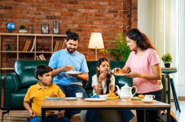 Hintli aile evde çayla samosa atıştırmalıkları yiyor, ebeveynler ve çocuklar sıcak oturma odasında oturmuş sohbet ederken, gülerken ve kaliteli bir bağ kurma zamanı geçirirken kızarmış yiyecek ve içeceklerin tadını çıkarıyorlar.