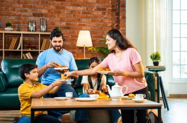 Hintli aile evde çayla samosa atıştırmalıkları yiyor, ebeveynler ve çocuklar sıcak oturma odasında oturmuş sohbet ederken, gülerken ve kaliteli bir bağ kurma zamanı geçirirken kızarmış yiyecek ve içeceklerin tadını çıkarıyorlar.