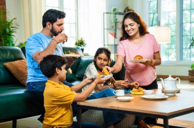 Hintli aile evde çayla samosa atıştırmalıkları yiyor, ebeveynler ve çocuklar sıcak oturma odasında oturmuş sohbet ederken, gülerken ve kaliteli bir bağ kurma zamanı geçirirken kızarmış yiyecek ve içeceklerin tadını çıkarıyorlar.
