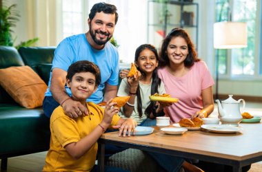 Hintli aile evde çayla samosa atıştırmalıkları yiyor, ebeveynler ve çocuklar sıcak oturma odasında oturmuş sohbet ederken, gülerken ve kaliteli bir bağ kurma zamanı geçirirken kızarmış yiyecek ve içeceklerin tadını çıkarıyorlar.