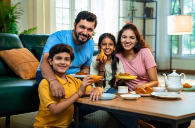 Hintli aile evde çayla samosa atıştırmalıkları yiyor, ebeveynler ve çocuklar sıcak oturma odasında oturmuş sohbet ederken, gülerken ve kaliteli bir bağ kurma zamanı geçirirken kızarmış yiyecek ve içeceklerin tadını çıkarıyorlar.