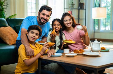 Hintli aile evde çayla samosa atıştırmalıkları yiyor, ebeveynler ve çocuklar sıcak oturma odasında oturmuş sohbet ederken, gülerken ve kaliteli bir bağ kurma zamanı geçirirken kızarmış yiyecek ve içeceklerin tadını çıkarıyorlar.