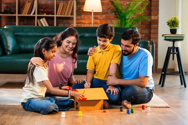 Hintli kardeşler karton ev modelini fırçayla boyarken ebeveynler evde neşe içinde oturup lüks oturma odasında oturup emlak hayallerini ve aile bağlarını sembolize ediyorlar.