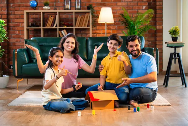 Hintli kardeşler karton ev modelini fırçayla boyarken ebeveynler evde neşe içinde oturup lüks oturma odasında oturup emlak hayallerini ve aile bağlarını sembolize ediyorlar.