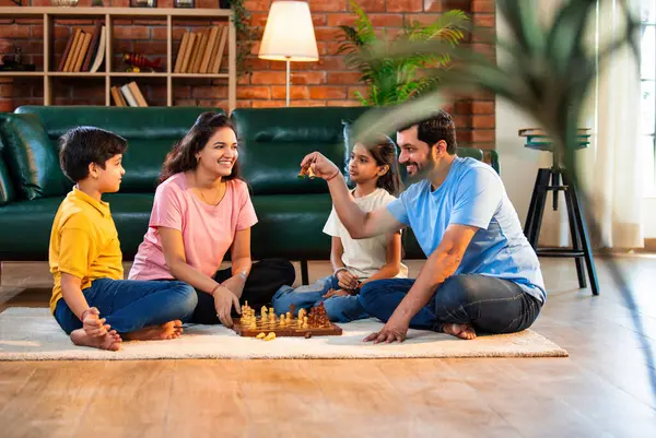Hintli aile satranç oynuyor. Halı döşemesinde şık bir oturma odasında oturuyorlar. Çocuklar ve ebeveynlerle evde vakit geçirip hafta sonu aktivitelerinin keyfini çıkarıyorlar.