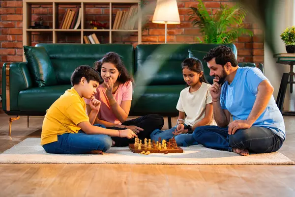Hintli aile satranç oynuyor. Halı döşemesinde şık bir oturma odasında oturuyorlar. Çocuklar ve ebeveynlerle evde vakit geçirip hafta sonu aktivitelerinin keyfini çıkarıyorlar.