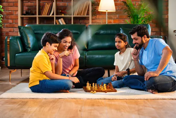 Hintli aile satranç oynuyor. Halı döşemesinde şık bir oturma odasında oturuyorlar. Çocuklar ve ebeveynlerle evde vakit geçirip hafta sonu aktivitelerinin keyfini çıkarıyorlar.