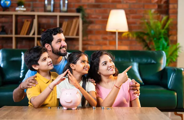 Çocuklar domuz kumbarasında para biriktirirken Hintli aileler arka planda gülümsüyor, mutlu oğul ve kız para kutusuna bozuk para koyuyorlar, evde rahat oturma odasında mali alışkanlıklar öğreniyorlar.