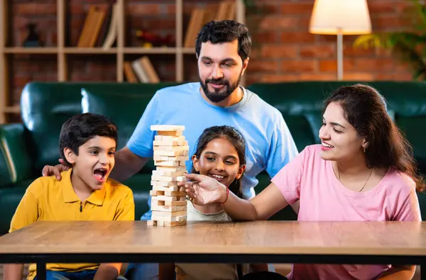 Hintli ebeveynler küçük çocuklarla vakit geçirmenin keyfini çıkarırken ve rahat modern oturma odasında ahşap blokları dikkatlice dengelerken aile evde ahşap blok oyunu oynuyor.