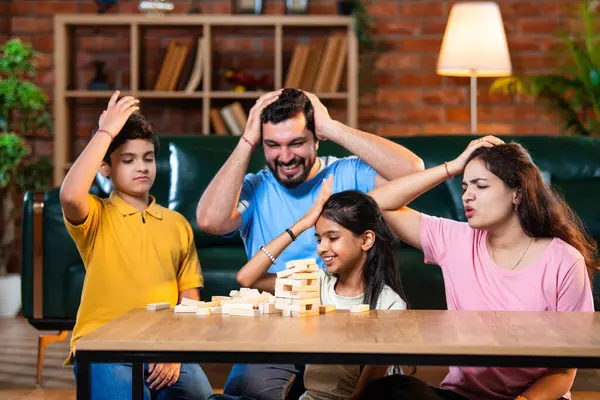 Hintli ebeveynler küçük çocuklarla vakit geçirmenin keyfini çıkarırken ve rahat modern oturma odasında ahşap blokları dikkatlice dengelerken aile evde ahşap blok oyunu oynuyor.
