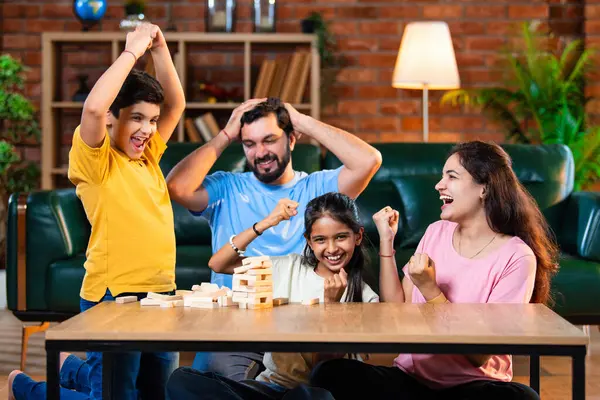 Hintli ebeveynler küçük çocuklarla vakit geçirmenin keyfini çıkarırken ve rahat modern oturma odasında ahşap blokları dikkatlice dengelerken aile evde ahşap blok oyunu oynuyor.