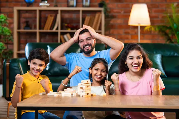 Hintli ebeveynler küçük çocuklarla vakit geçirmenin keyfini çıkarırken ve rahat modern oturma odasında ahşap blokları dikkatlice dengelerken aile evde ahşap blok oyunu oynuyor.