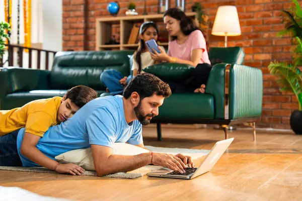 Baba oğul çalışırken Hintli adam dizüstü bilgisayarı yerde yatarken ve çocuğu üzerinde kestirirken, anne ve kızı rahat modern oturma odasında akıllı telefon kullanarak koltukta otururken, baba çalışıyor.