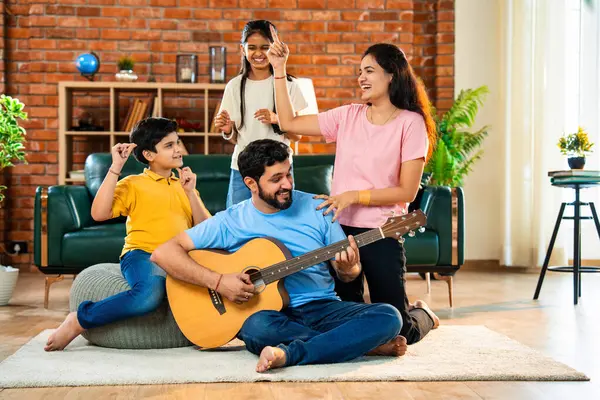 Babam gülen Hintli eş, oğul ve kızıyla gitar çalıyor. Müsrif oturma odasında oturuyor. Müzikal kaynaşmanın ve modern evde kaliteli zaman geçirmenin tadını çıkarıyor.