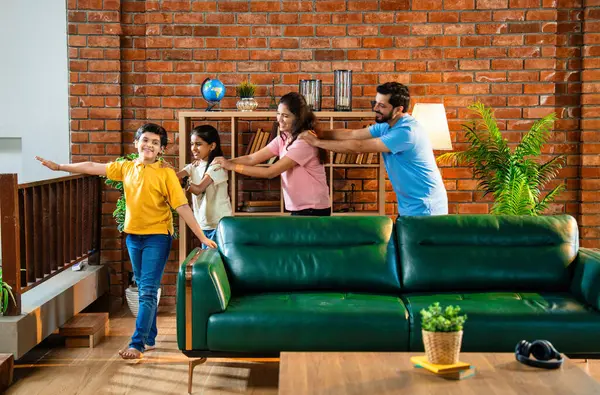 Mutlu Hintli aile içeride tren oyunu oynuyor, küçük çocuk motor gibi yol gösteriyor, ebeveynler de onu takip ediyor, evde rahat bir oturma odasında birlikte koşarken eğleniyorlar.