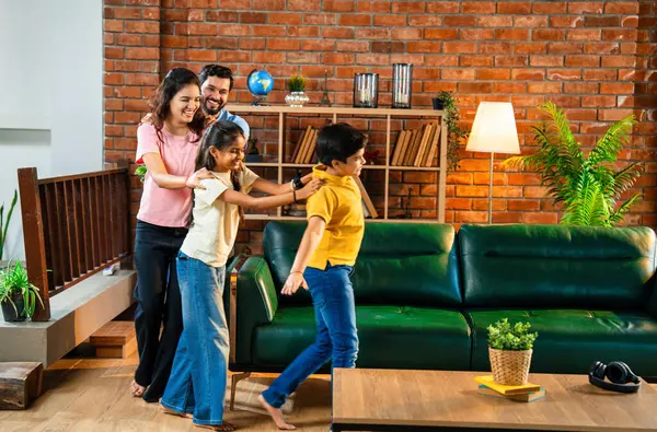 Mutlu Hintli aile içeride tren oyunu oynuyor, küçük çocuk motor gibi yol gösteriyor, ebeveynler de onu takip ediyor, evde rahat bir oturma odasında birlikte koşarken eğleniyorlar.