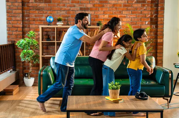 Mutlu Hintli aile içeride tren oyunu oynuyor, küçük çocuk motor gibi yol gösteriyor, ebeveynler de onu takip ediyor, evde rahat bir oturma odasında birlikte koşarken eğleniyorlar.