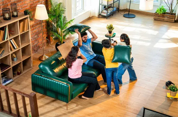 Hintli aile evde yastık savaşı yapıyor, aileler ve çocuklar rahat bir oturma odasında gülüyor ve eğlenirken eğlenceli bağlılık anlarının tadını çıkarıyor, evde neşe ve kaliteli aile zamanlarını paylaşıyorlar.