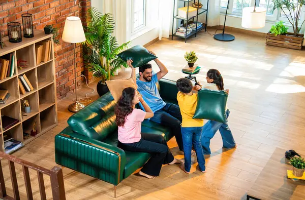 Hintli aile evde yastık savaşı yapıyor, aileler ve çocuklar rahat bir oturma odasında gülüyor ve eğlenirken eğlenceli bağlılık anlarının tadını çıkarıyor, evde neşe ve kaliteli aile zamanlarını paylaşıyorlar.