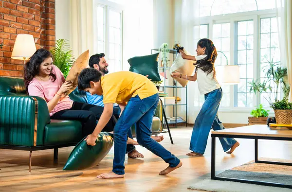 Hintli aile evde yastık savaşı yapıyor, aileler ve çocuklar rahat bir oturma odasında gülüyor ve eğlenirken eğlenceli bağlılık anlarının tadını çıkarıyor, evde neşe ve kaliteli aile zamanlarını paylaşıyorlar.