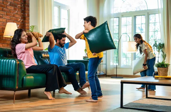 Hintli aile evde yastık savaşı yapıyor, aileler ve çocuklar rahat bir oturma odasında gülüyor ve eğlenirken eğlenceli bağlılık anlarının tadını çıkarıyor, evde neşe ve kaliteli aile zamanlarını paylaşıyorlar.