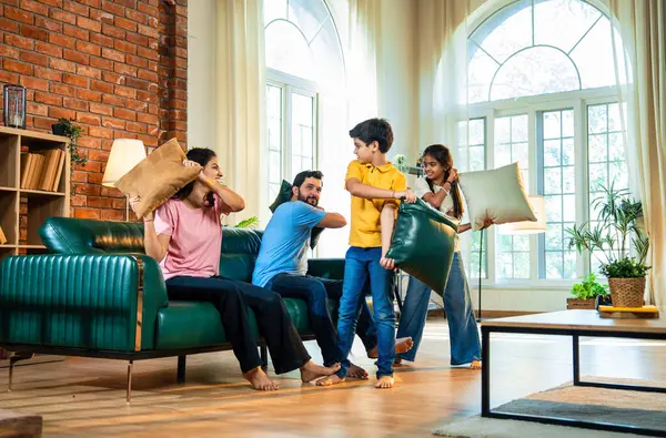 Hintli aile evde yastık savaşı yapıyor, aileler ve çocuklar rahat bir oturma odasında gülüyor ve eğlenirken eğlenceli bağlılık anlarının tadını çıkarıyor, evde neşe ve kaliteli aile zamanlarını paylaşıyorlar.