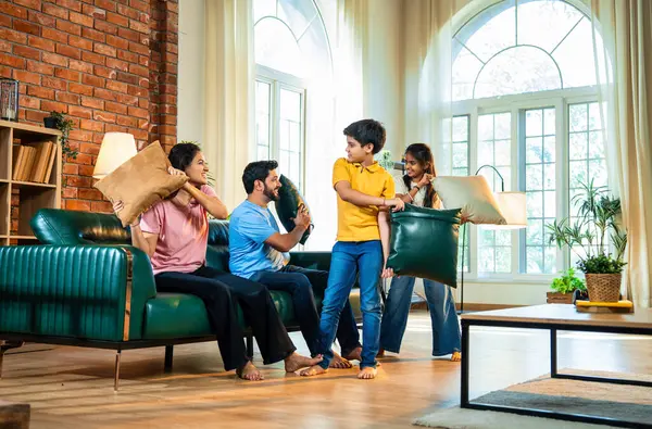Hintli aile evde yastık savaşı yapıyor, aileler ve çocuklar rahat bir oturma odasında gülüyor ve eğlenirken eğlenceli bağlılık anlarının tadını çıkarıyor, evde neşe ve kaliteli aile zamanlarını paylaşıyorlar.