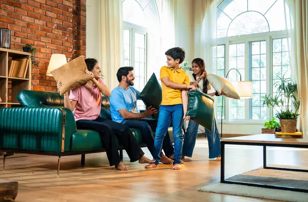 Hintli aile evde yastık savaşı yapıyor, aileler ve çocuklar rahat bir oturma odasında gülüyor ve eğlenirken eğlenceli bağlılık anlarının tadını çıkarıyor, evde neşe ve kaliteli aile zamanlarını paylaşıyorlar.