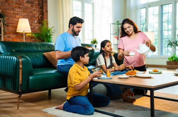 Hintli aile evde çayla samosa atıştırmalıkları yiyor, ebeveynler ve çocuklar sıcak oturma odasında oturmuş sohbet ederken, gülerken ve kaliteli bir bağ kurma zamanı geçirirken kızarmış yiyecek ve içeceklerin tadını çıkarıyorlar.