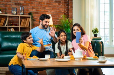 Evde samosa yiyip çay içerken görüntülü konuşma, çocuklu Hintli aileler arkadaşlarıyla ya da akrabalarıyla sohbet etme, rahat modern oturma odasında atıştırmalıkların tadını çıkarma.