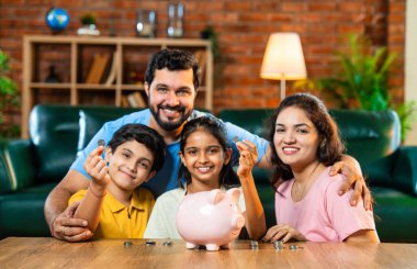 Çocuklar domuz kumbarasında para biriktirirken Hintli aileler arka planda gülümsüyor, mutlu oğul ve kız para kutusuna bozuk para koyuyorlar, evde rahat oturma odasında mali alışkanlıklar öğreniyorlar.