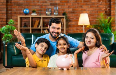 Çocuklar domuz kumbarasında para biriktirirken Hintli aileler arka planda gülümsüyor, mutlu oğul ve kız para kutusuna bozuk para koyuyorlar, evde rahat oturma odasında mali alışkanlıklar öğreniyorlar.