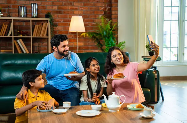 Evde samosa yiyip çay içerken selfie çekmek, iki çocuklu mutlu Hintli ebeveynler atıştırmalıkların tadını çıkarmak, neşeli yakınlaşma anları yakalamak ve modern sıcak oturma odasında birlikte vakit geçirmek.
