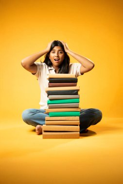 Kitap Yığını 'yla hüzünle oturan, Gergin Hintli Genç Kız Sarı Kusursuz Arkaplanda Bunalmış, Çalışmaların ve Etrafındaki Eğitim Kitapları Yığını ile Akademik Baskının Yükünü Hissediyor
