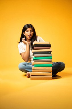 Kitap Yığını 'yla hüzünle oturan, Gergin Hintli Genç Kız Sarı Kusursuz Arkaplanda Bunalmış, Çalışmaların ve Etrafındaki Eğitim Kitapları Yığını ile Akademik Baskının Yükünü Hissediyor