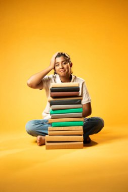 Kitap Yığını 'yla hüzünle oturan, Gergin Hintli Genç Kız Sarı Kusursuz Arkaplanda Bunalmış, Çalışmaların ve Etrafındaki Eğitim Kitapları Yığını ile Akademik Baskının Yükünü Hissediyor